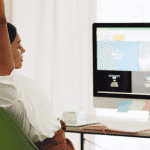 A person stretching in an office chair with hands raised, in front of a computer displaying a website design, in a bright and plant-decorated office environment.