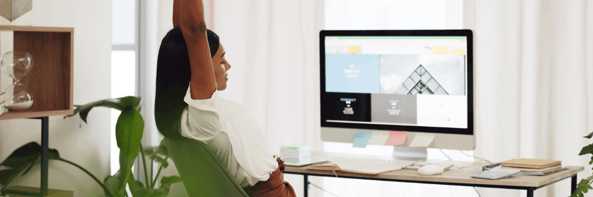 A person stretching in an office chair with hands raised, in front of a computer displaying a website design, in a bright and plant-decorated office environment.